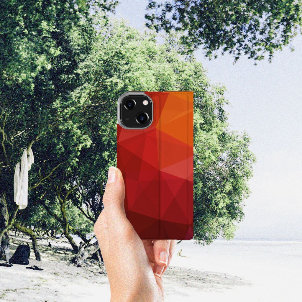 Stand Case voor iPhone 14 Polygon Red in hand, op een zonnige strandlocatie met groene bomen.