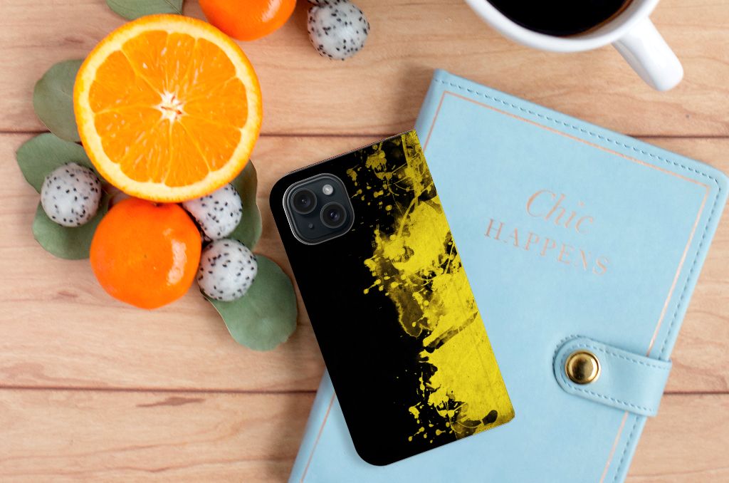 iPhone 15 Standcase België met zwart-geel design op een houten tafel met fruit en een notitieboekje
