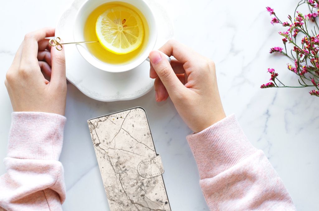 Samsung Galaxy A14 5G Bookcase Marmer Beige met een hand boven een kopje thee en citroen, op een marmeren tafel.
