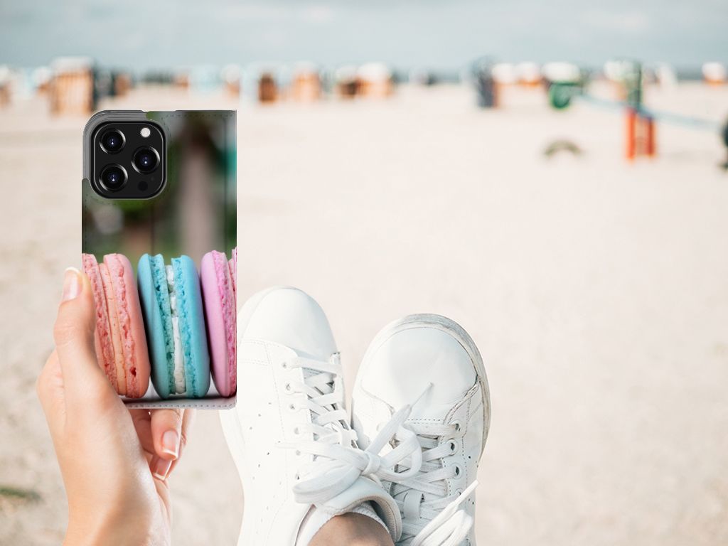 Vrouw houdt een iPhone 13 Pro Flip Style Cover Macarons vast met kleurige macarons, op een strandachtergrond.