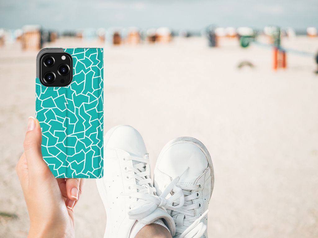 Hand holding an iPhone 13 Pro Bookcase Cracks Blue design at the beach with white sneakers.
