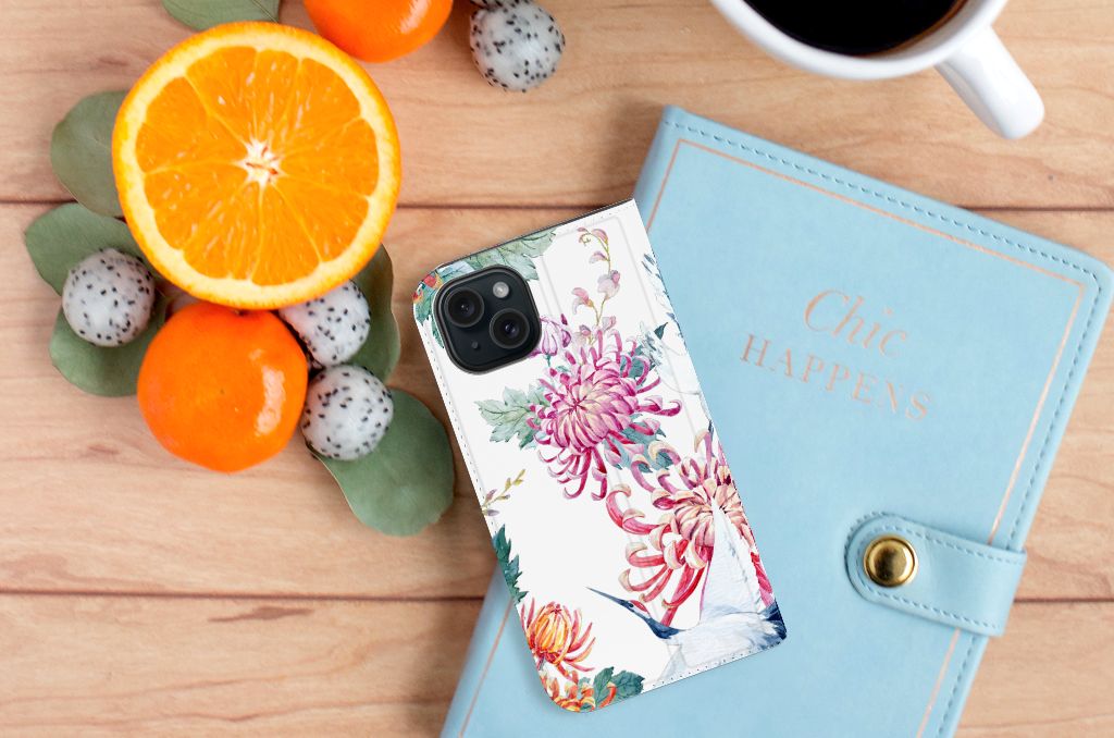 iPhone 15 Hoesje maken Bird Flowers met bloemen en ooievaars, omringd door fruit en een notitieboek.