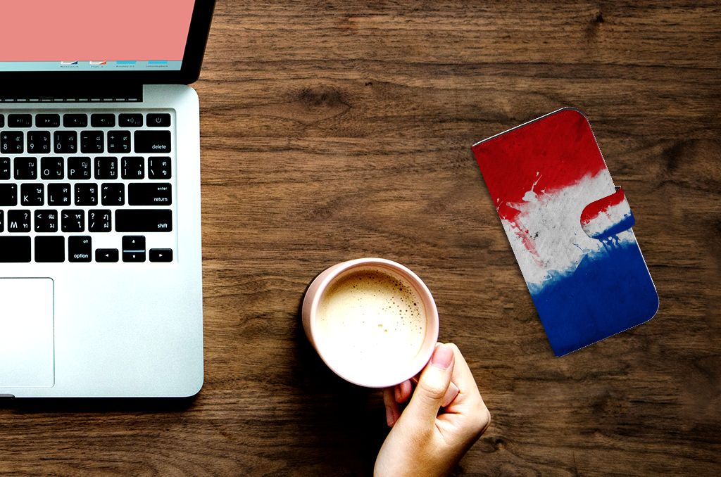 Hand holding coffee cup beside iPhone 15 Pro Bookstyle Case Nederland on a wooden table with a laptop.