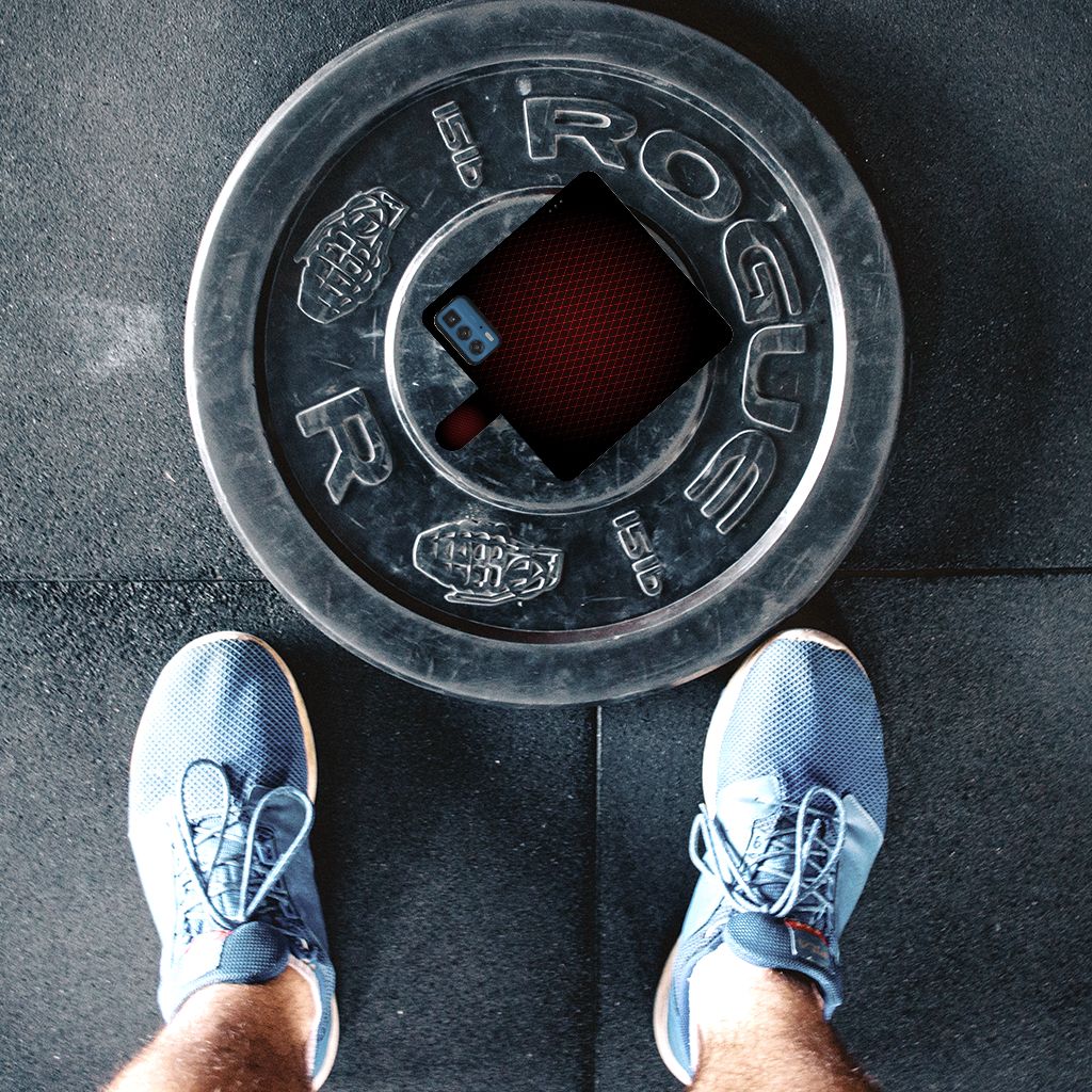 Rogue gewichtschijf met smartphone in het midden en sportieve schoenen in de voorgrond op een gymvloer.