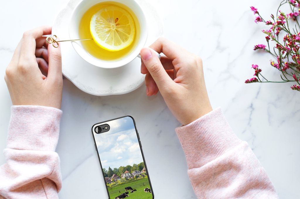 Hand holding a cup of tea next to a colorful iPhone SE 2022 | SE 2020 | 7/8 Dierenprint Telefoonhoesje Koeien on a table.