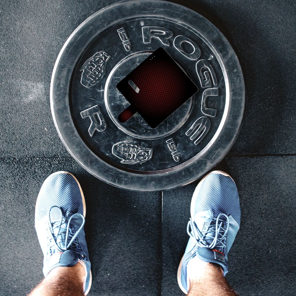 Perspectief van schoenen boven een Rogue gewichtsschijf met zwarte en rode details op een fitnessvloer.