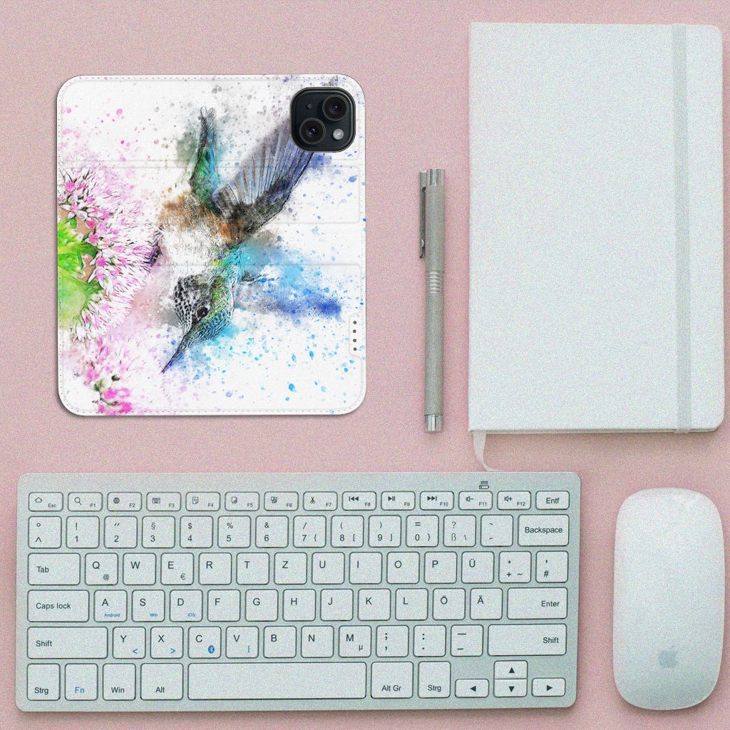Bookcase iPhone 15 Vogel met kleurrijke vogelillustratie en bloemen op een bureau met toetsenbord en notitieboekje.