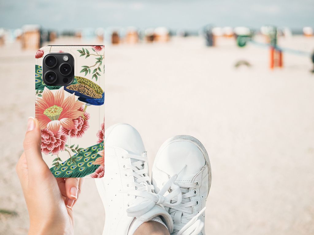 Hoesje maken voor iPhone 15 Pro Pink Peacock met bloemenontwerp en pauwen, vastgehouden op strand.