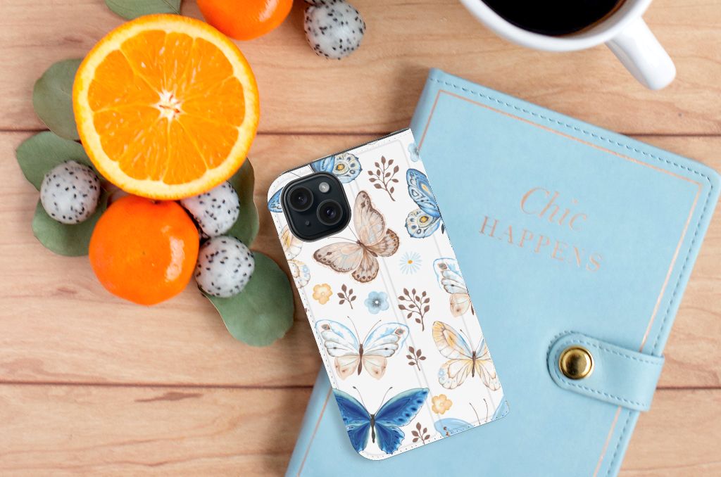 Hoesje maken voor iPhone 15 Vlinder met kleurrijke vlinders op een vlakke ondergrond met fruit en een notitieboekje.