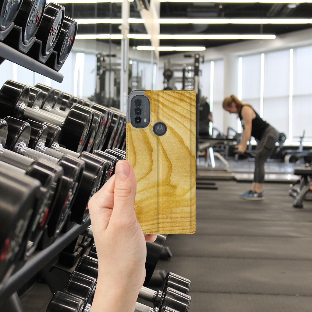 Motorola Moto E20 Book Wallet Case Licht Hout hand vast houdend in sportschool met gewichten op de achtergrond