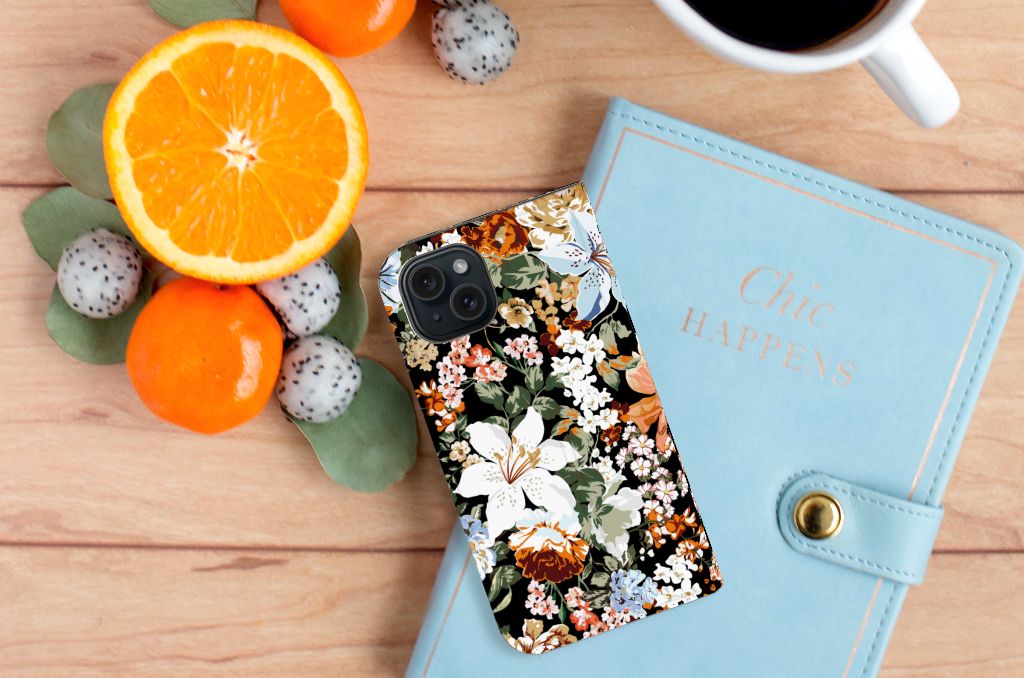 Smart Cover voor iPhone 15 Dark Flowers met bloemenprint op een houten tafel naast fruit en een notitieboekje.