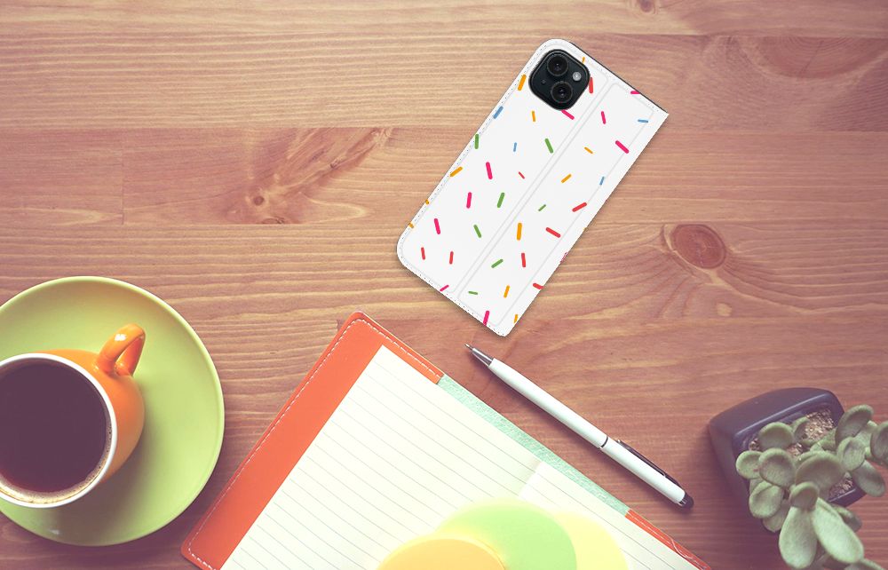 iPhone 15 Plus Flip Style Cover Donut Roze met kleurrijke sprinkles op een houten tafel met koffie en notitieboek.