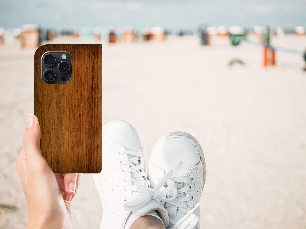 Hand die een iPhone 15 Pro Book Wallet Case Donker Hout vasthoudt in een strandomgeving.