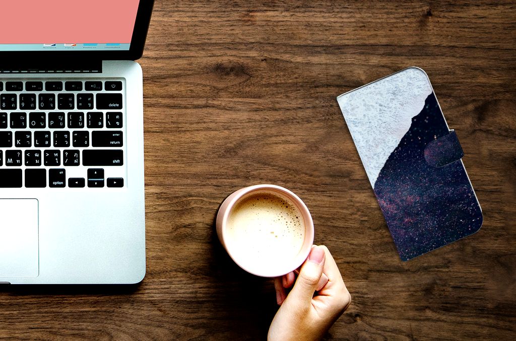 Hand holding a cup of coffee beside a laptop with an iPhone 15 Pro Max Book Case Sea in Space on the table.